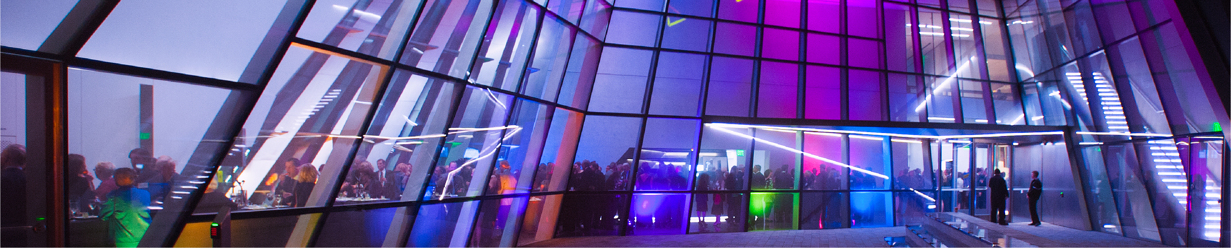 The MSU Broad Art Museum at night during a gala event. The museum is lit by different blue, purple, and pink lights. People can be seen indoors.
